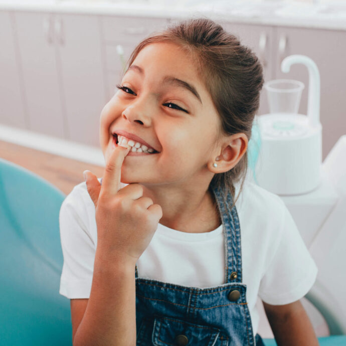 girl sitting in a dentist chair happily pointing to her teeth